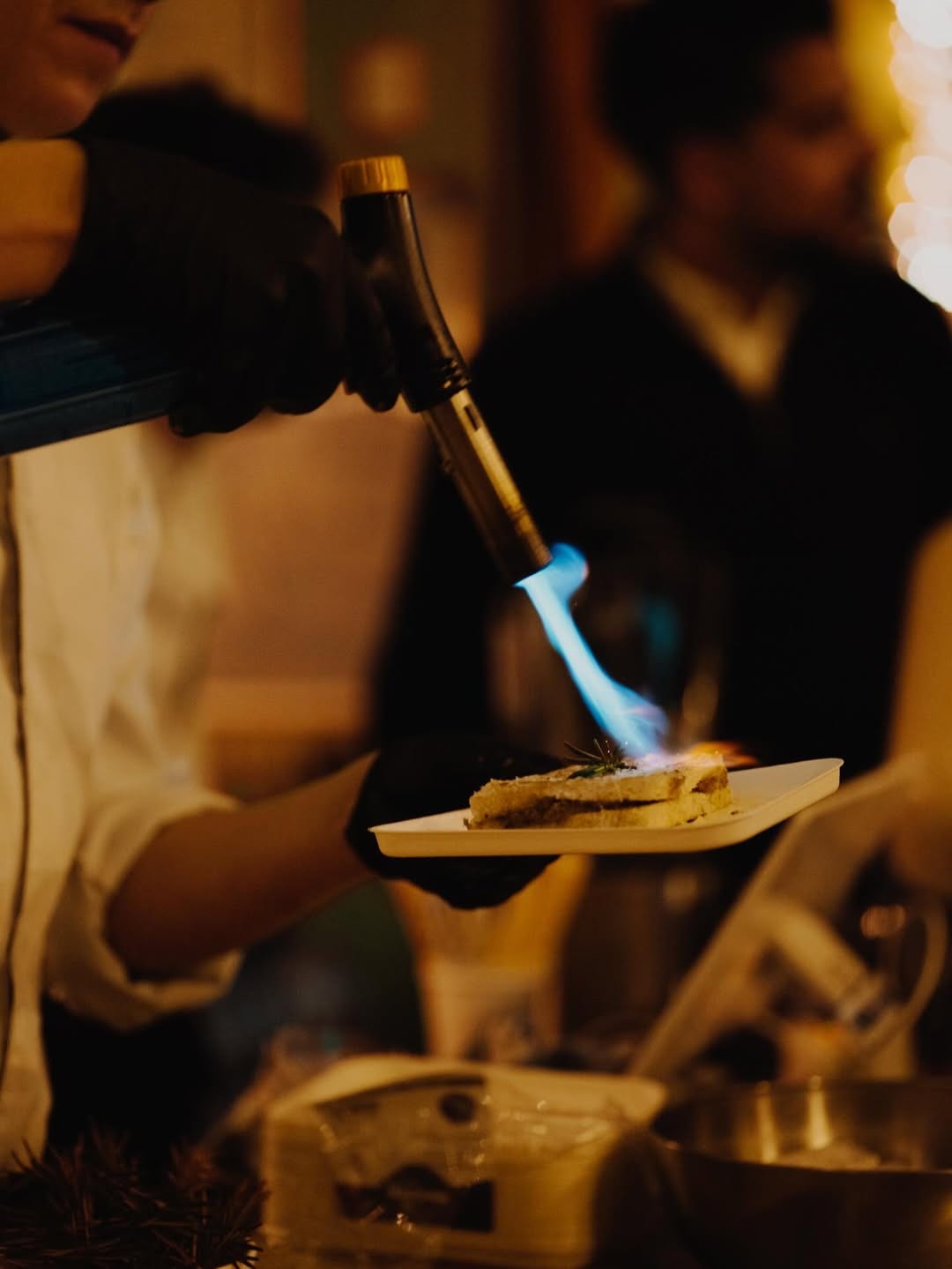 A chef torching a brioche montadito to finish at an El Payo night — blue flame, warm event light.